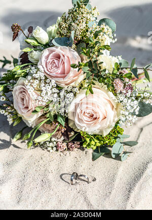 Schöne Hochzeit Ringe liegen in den Sand am Strand vor dem Hintergrund einer Braut Blumenstrauß. Stockfoto