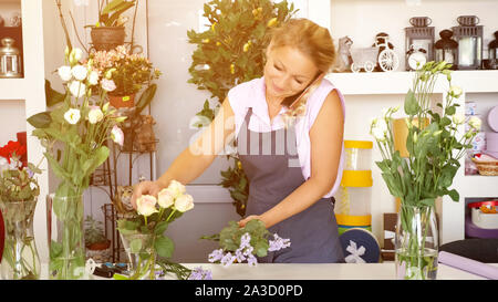 Professionelle Frau florist Gespräche auf dem Smartphone, um und macht Strauß in Flower Shop arbeiten. Reife Frau in Schürze im Bereich Floristik studio schöne Bündel, Sonnenlicht Stockfoto