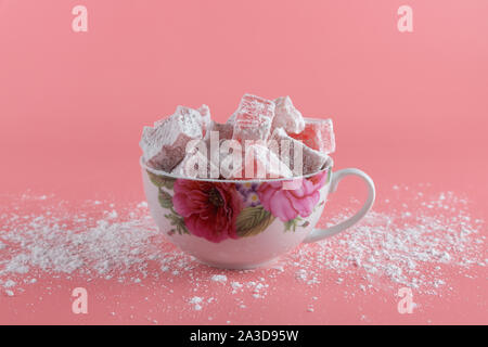 Eine weiße Tasse mit gemalten Blumen voller Lokum, ein Türkischer Honig, auf einem rosa Hintergrund und Puderzucker auf dem Tisch verschüttet Stockfoto