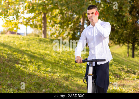 Freudige und legerer Mann reiten Elektroroller im Stadtpark und am Telefon sprechen Stockfoto