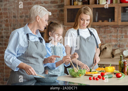 Süße kleine Mädchen mischen Salat unter Mama und Oma Aufsicht Stockfoto