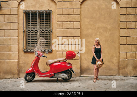 Attraktive junge Frau mit Hut in der Hand durch die alten Gebäude aus Stein stehen und geparkten roten Motorrad am Gehsteig in der Toskana Italien Stockfoto