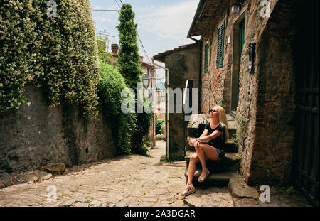 Sorglos Frau in Sonnenbrille und casual Outfit auf Schritte durch Eingang der alten Backsteinhaus in der Toskana Italien sitzen Stockfoto