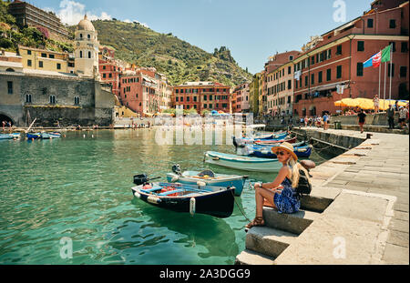 Seitenansicht der glücklichen Frau in Hut und Sonnenbrille sitzt am Pier in der Nähe günstig bunte Boote an ländliche Stadt mit alten Gebäuden im Sommer Tag Stockfoto