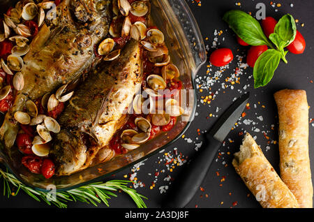 Dorade mit Muscheln in Tomaten mit Olivenöl im Ofen gebackene Brot auf schwarzem Hintergrund. Mediterrane Küche Konzept Stockfoto
