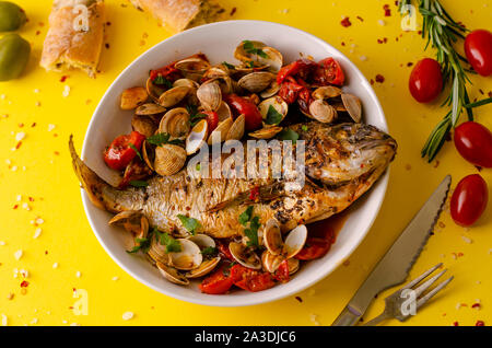 Gebackene dorada Fisch mit Muscheln in einer weißen Schüssel auf gelben Hintergrund. Meeresfrüchte Konzept. Overhead shot. Stockfoto