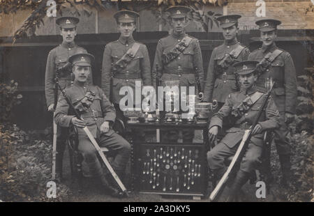 Jahrgang 1914 Photographische Postkarte zeigt eine Gruppe von WW1 British Army Soldaten mit ihren Gewehren. Stolz Sitzen und Stehen mit ihren Trophäen, Pokale und Silber Löffel Stockfoto