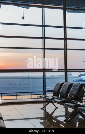 Blick auf Flughafen, interoir leeren Bank Stühle in der Abflughalle bei Sonnenaufgang. Flugzeug und Gebäude im Hintergrund. Reise und Verkehr Konzept Stockfoto