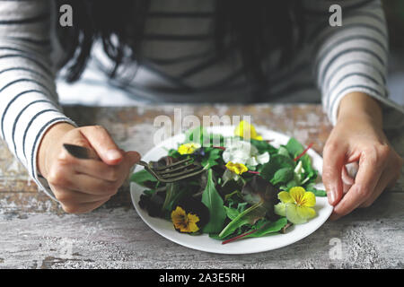 Selektive konzentrieren. Eine schöne Mischung aus Salaten mit Blumen auf einem weißen Teller wird von einem Mädchen statt. Diät Konzept. Gesund Vegan essen. Stockfoto