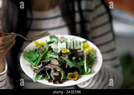 Selektive konzentrieren. Eine schöne Mischung aus Salaten mit Blumen auf einem weißen Teller wird von einem Mädchen statt. Diät Konzept. Gesund Vegan essen. Stockfoto