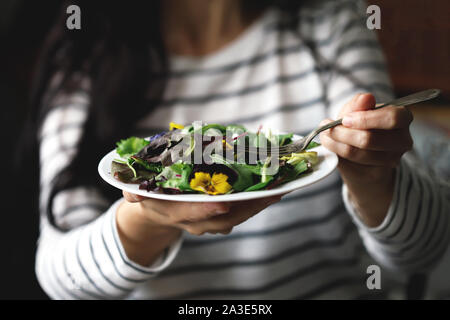 Selektive konzentrieren. Eine schöne Mischung aus Salaten mit Blumen auf einem weißen Teller wird von einem Mädchen statt. Diät Konzept. Gesund Vegan essen. Stockfoto