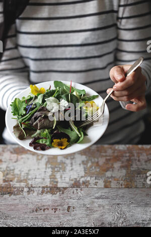 Selektive konzentrieren. Eine schöne Mischung aus Salaten mit Blumen auf einem weißen Teller wird von einem Mädchen statt. Diät Konzept. Gesund Vegan essen. Stockfoto