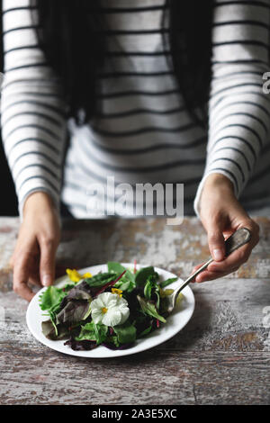 Selektive konzentrieren. Eine schöne Mischung aus Salaten mit Blumen auf einem weißen Teller wird von einem Mädchen statt. Diät Konzept. Gesund Vegan essen. Stockfoto