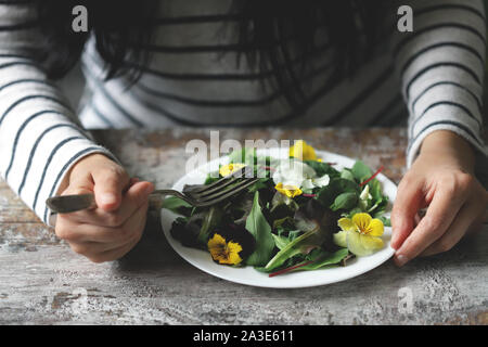 Selektive konzentrieren. Eine schöne Mischung aus Salaten mit Blumen auf einem weißen Teller wird von einem Mädchen statt. Diät Konzept. Gesund Vegan essen. Stockfoto