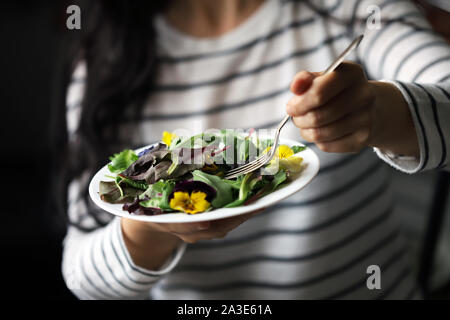 Selektive konzentrieren. Eine schöne Mischung aus Salaten mit Blumen auf einem weißen Teller wird von einem Mädchen statt. Diät Konzept. Gesund Vegan essen. Stockfoto