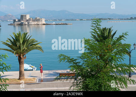 Nafplio, Griechenland - 19 Juli 2019; Frau in rot gemusterten Drees steht über Handy in Aussicht auf die promenade zu Insel Bourtzi mit alten Festung se Stockfoto