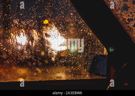 Nacht Stadt bokeh Lichter durch gefrorene Auto Fenster. Blurry auto Silhouette durch geschmolzenen Schnee und Wassertropfen auf der Windschutzscheibe Auto gesehen. Hellen bunten Abend Winter. Stockfoto