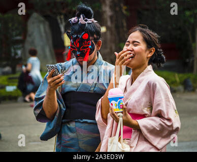 Eine rot-schwarze Kitsune-Fuchsmaske bedeckt das Gesicht eines Mannes in einem Kimono, der mit einer lachenden Frau in Asakusa im Taito-Bezirk von Tokio, Japan, ein Selfie macht. Die Frau hält eine Tasse Kakigori-Geschabtes Eis. Stockfoto