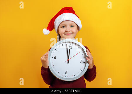 Heiligabend. Portrait Of Happy charmante kleine süße Mädchen in Santa's Hut. Das Kind hält eine Uhr auf einem gelben Hintergrund. Stockfoto