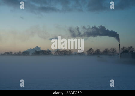 Sonnenuntergang an einem kalten Wintertag, mit einem rauchenden Schornstein. Die Sonne hinter Wolken, berühren den Horizont hinter einer Reihe von kleinen Bäumen. Stockfoto