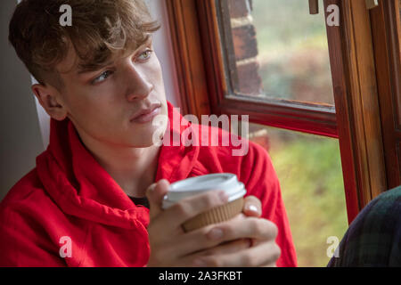 Traurig, deprimiert nachdenklich jugendlich Junge männliche Teenager jungen Mann, der aus einem Fenster trinken Kaffee oder Tee suchen Stockfoto