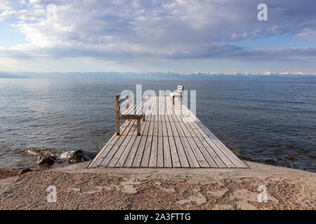 Pier mit Bänken am Issyk-Kul See mit einer Bergkette mit schneebedeckten Gipfel am Horizont gegen einen bewölkten Himmel Stockfoto