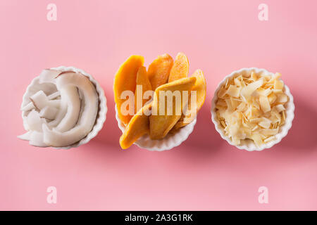 Entwässert und getrocknet Mango und Kokos Chips in weiße Schüssel auf rosa Hintergrund. Blick von oben. Stockfoto
