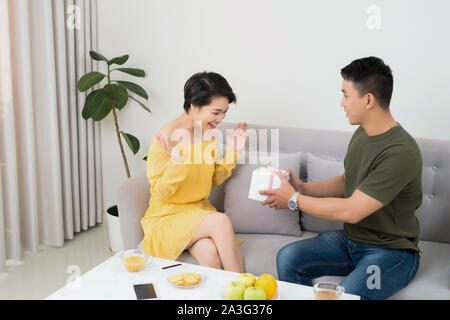 Mann überrascht, seine Freundin mit einem Geschenk auf der Couch zu Hause im Wohnzimmer Stockfoto