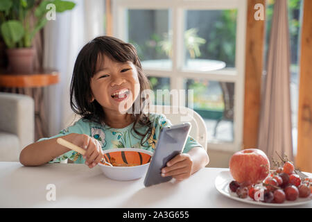 Glückliche junge Kleinkind ihr Frühstück genießen, während Sie smartphone Stockfoto