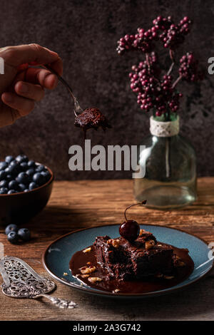 Kaukasische männliche Hand, denn ein Stück vegane Schokolade Brownie mit Gebäck Gabel, Schokoladensauce, tropft unten auf einem feuchten brownie Stockfoto
