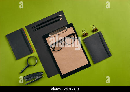 Blatt schwarzes Papier, Zwischenablage mit braunem Papier, Lupe, Gläser, Hefter, Bleistifte, zwei Büroklammern und Ordner auf grünem Hintergrund mit Kopie spac Stockfoto