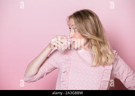 Junge schwangere Frau mit rosa Kleid Trinkwasser oder Medikation aus Plastikbecher. Studio shot auf rosa Hintergrund. Flüssigkeitszufuhr während der Schwangerschaft conc Stockfoto