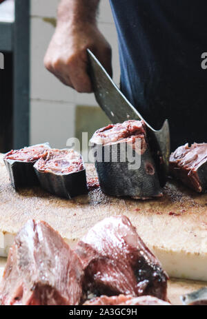 Detail von einem Fischer schneidet Thunfisch mit einem riesigen Cleaver auf einem Holzbrett. Verschwommen Thunfisch, Fleisch in den Vordergrund. Traditionellen Fischmarkt in Funchal, Madeira, Portugal. Fischerei, Fischerei. Stockfoto