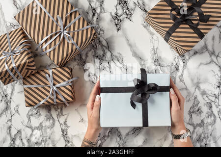 Hände einer Frau halten Weißbuch Geschenkkarton mit schwarzen Band und Bogen gegen einen Marmor Hintergrund gebunden. Gestreift Braun vorhanden sind Festlegung der n Stockfoto