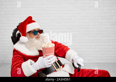 Seitenansicht der entspannte Weihnachtsmann mit grauen Bart tragen Tracht und 3D-Brille, sitzen bequem im Sessel und halten Schaufel mit Popcorn in der einen Hand und TV-Zapper in eine andere Hand. Stockfoto