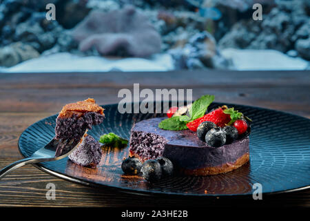 Selektiver Fokus der leckere blueberry Cheesecake mit Erdbeeren, roten Beeren und Minze auf schwarze Platte im Restaurant serviert. Gabel mit Stück süße Nachspeise. Konzept der Creme und Essen. Stockfoto