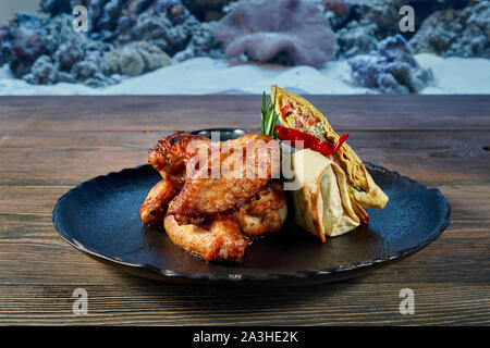 Ansicht von der Seite der leckeren Brötchen mit Gemüse und Käse und Saftig gegrillte Chicken Wings auf schwarzem Teller serviert. Leckeres Essen, die auf der Tabelle auf der verschwommenen Hintergrund. Konzept der Speisen und Teller. Stockfoto