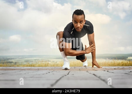 Schöner Athlet Übungen, Stretching. Mann mit sportlicher Körper tragen in schwarzen T-Shirt und Shorts Training am Morgen. Sportsman posiert im Freien, an der Kamera schaut. Stockfoto