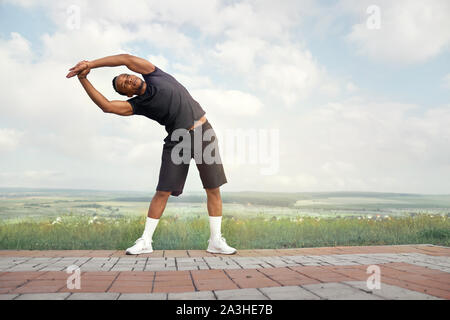 Stattliche Sportler, die Pisten im Freien. Aktiven Sportler mit muskulösen Körper in schwarzen T-Shirt und schwarze Shorts Übungen. Athletischer Mann die Hände, auf dem Hintergrund des blauen Himmels posieren. Stockfoto