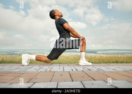 Muskulöse Athleten mit in schwarzen T-Shirt, Shorts und weißen Socken, Sneakers, posiert im Freien Seitenansicht. Schöner mann Übungen mit Stretching für die Füße am Morgen. Stockfoto