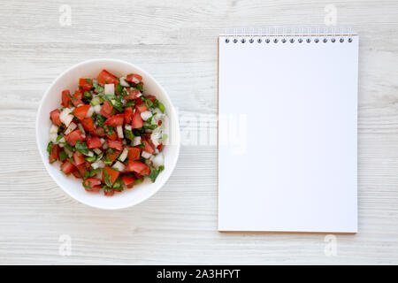 Pico de Gallo in einer weißen Schüssel, leere Notepad auf weißem Holz- Oberfläche, Ansicht von oben. Overhead, von oben, flach. Kopieren Sie Platz. Stockfoto