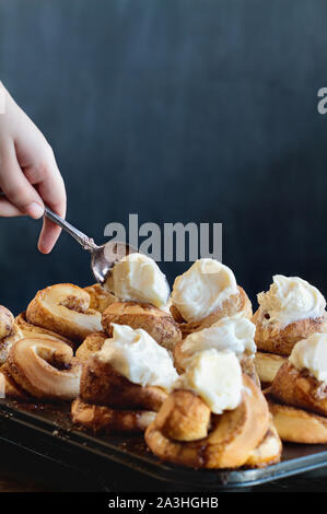 Woman's Hand dekorieren frischen hausgemachten hohen Zimt Brötchen mit Frischkäse Puderzucker. Selektiver Fokus mit unscharfen Vordergrund und Hintergrund. Stockfoto