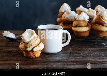 Frischen hausgemachten hohen Cinnamon Roll mit Frischkäse Puderzucker. Heiß dampfende Tasse Kaffee im Hintergrund mit mehr Brötchen Kühlung auf Bäcker Zahnstange. Stockfoto