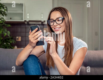 Hübsche brünette Frau mit Smartphone sitzt auf einem Sofa, drinnen geschossen Stockfoto