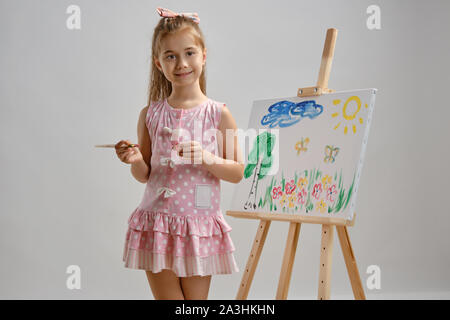 Kleines Mädchen Künstler in einem rosa Kleid steht hinter Staffelei und malen Mit Pinsel auf Leinwand, auf weissem studio Hintergrund isoliert. Medium close-up. Stockfoto