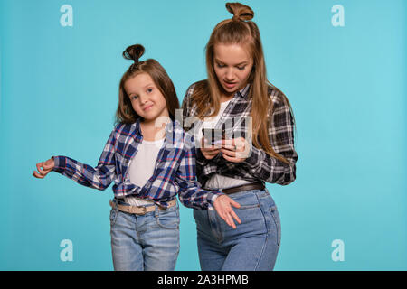 Mutter und Tochter in karierten Hemden und Blue Denim Jeans gekleidet sind Sie ihr Smartphone während vor einem blauen studio Hintergrund posiert. Nahaufnahme. Stockfoto