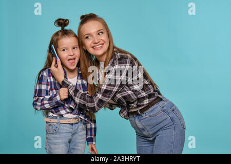 Mutter und Tochter in karierten Hemden und Blue Denim Jeans gekleidet sind Sie ihr Smartphone während vor einem blauen studio Hintergrund posiert. Nahaufnahme. Stockfoto