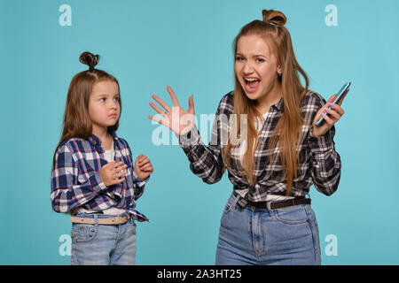 Mutter und Tochter in karierten Hemden und Blue Denim Jeans gekleidet sind Sie ihr Smartphone während vor einem blauen studio Hintergrund posiert. Nahaufnahme. Stockfoto