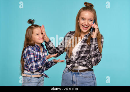 Mutter und Tochter in karierten Hemden und Blue Denim Jeans gekleidet sind Sie ihr Smartphone während vor einem blauen studio Hintergrund posiert. Nahaufnahme. Stockfoto