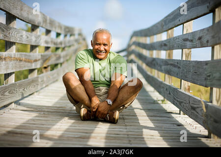 Porträt eines lächelnden reifen erwachsenen Mann Sitzen mit gekreuzten Beinen auf einem hölzernen Weg. Stockfoto